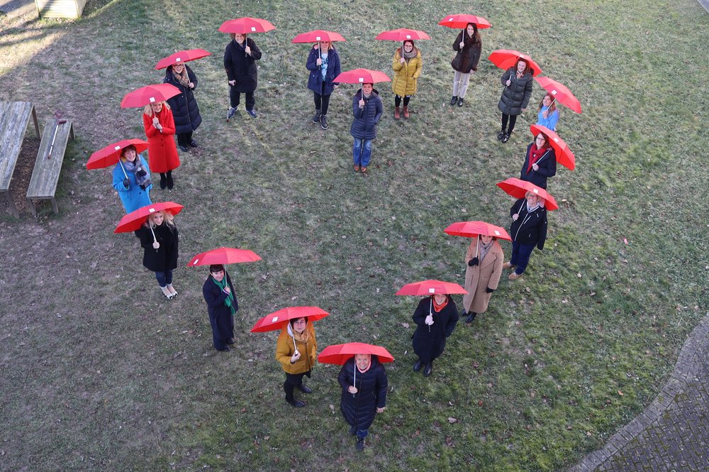 Knapp 20 Frauen haben sich in Form eines Herzens aufgestellt und halten einen roten Regenschirm in die Luft.