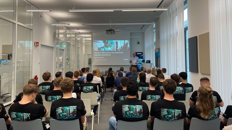 Gruppenbild: Teilnehmende des Hackathon Siemens sitzen in einem Raum und schauen auf eine große Leinwand.