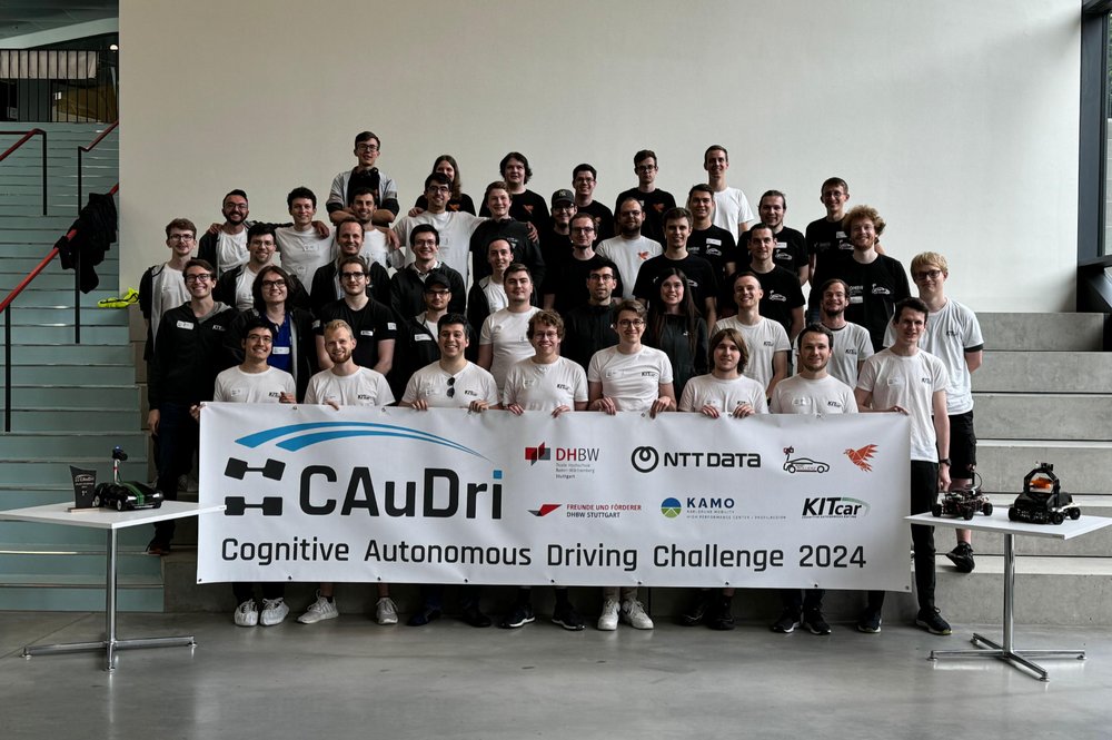 Die Teilnehmenden der CAuDri-Challenge 2024 - Gruppenfoto im Neubau Technik. Die Teilnehmenden halten ein Banner mit der Aufschrift Caudri-Cup vor sich. 