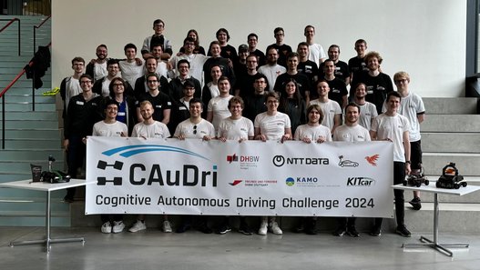 Die Teilnehmenden der CAuDri-Challenge 2024 - Gruppenfoto im Neubau Technik. Die Teilnehmenden halten ein Banner mit der Aufschrift Caudri-Cup vor sich. 