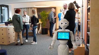 Roboter Pepper steht im Vordergrund, um die Gäste zu begrüßen, im Hintergrund informieren sich Besucher*innen über das Studio41.