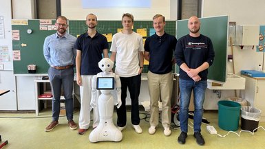 Gruppenbild mit dem Roboter Pepper im Klassenzimmer vor der Tafel.