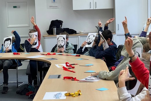 Das Zentrum für Managementsimulation (ZMS) spielte mit Schülerinnen und Schülern der fünften und sechsten Klasse das Planspiel „Fischteich“. Schüler*innen halten sich Papiermasken vor das Gesicht. 