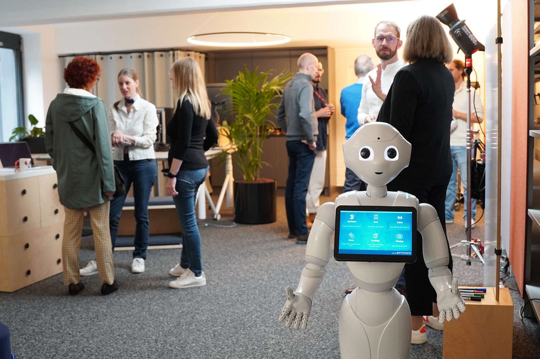 Im Vordergrund steht der humanoide Roboter Pepper, während im Hintergrund Gespräche stattfinden.